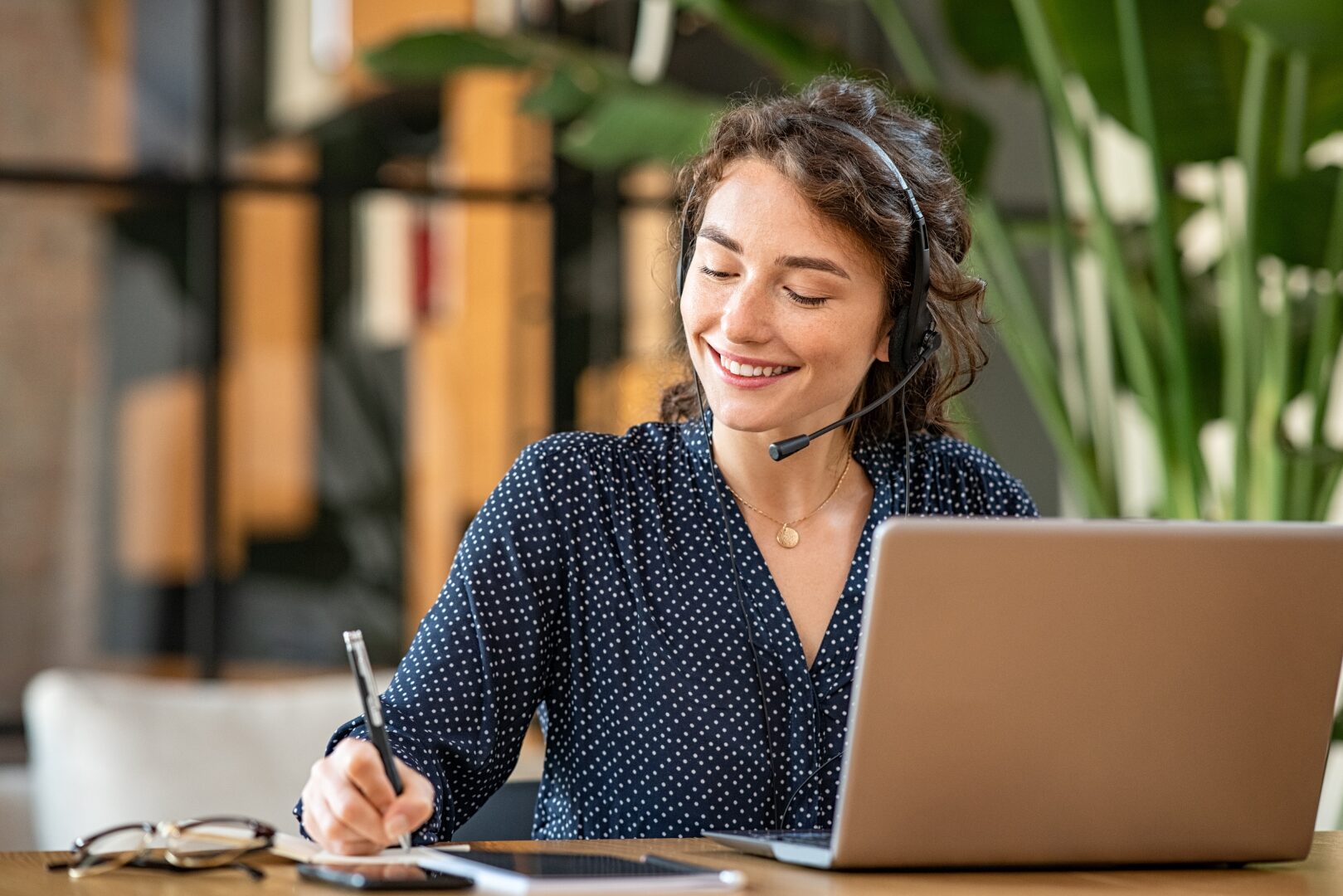 Mulher sentada em frente ao computador usando um fone de ouvido estilo headset sorrindo e anotando algo em um caderno, como se estivesse realizando anotações de uma reunião online.